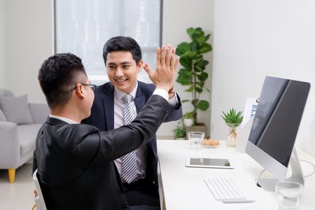 Two cheerful excited young men giving high five and working in office togetherの写真素材