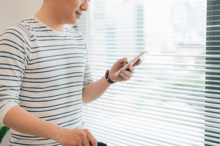 Smiling handsome young man using phone by the windowの写真素材