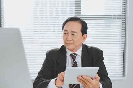 Senior (mature) businessman sitting at office in front of laptop and holding a digital tablet.の写真素材