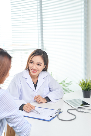 Female doctor talks to female patient in hospital office while writing on the patients health record on the table. Healthcare and medical service.の写真素材