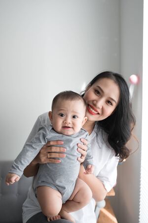 Beautiful young mom is playing with her cute baby and smiling while standing near the window at home