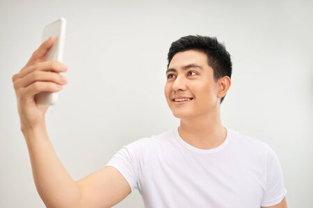 Smiling young asian man making selfie photo on smartphone over white background