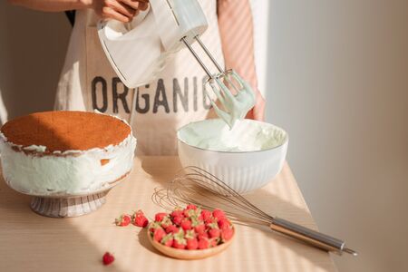 Male hands whipping whites cream in glass bowl with mixer on wooden table. Making sponge cake or red velvet cakeの写真素材