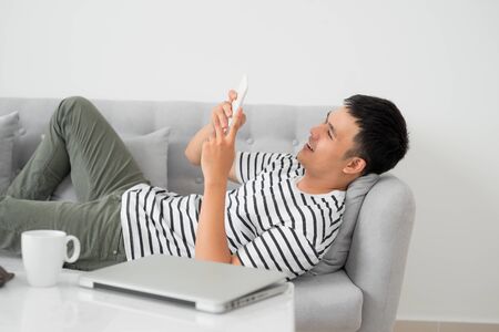 Asian young man using mobile phone while lying indoor.の写真素材