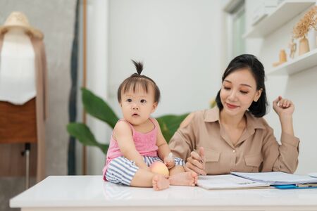 Little baby girl looking at camera while sitting on office desk with her mother in officeの写真素材