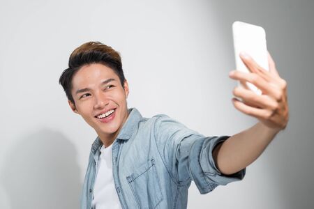 Studio shot of a smiling Asian young man making selfie with the smartphone while standing over white backgroundの写真素材