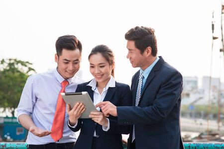 Asian businessman in suit showing online presentation on digital tablet to his colleagues while they standingの写真素材