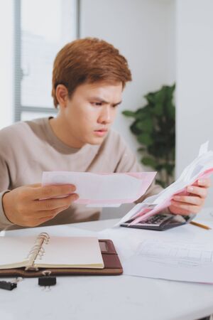Unhappy asian man looking at bills sitting at table in living roomの写真素材