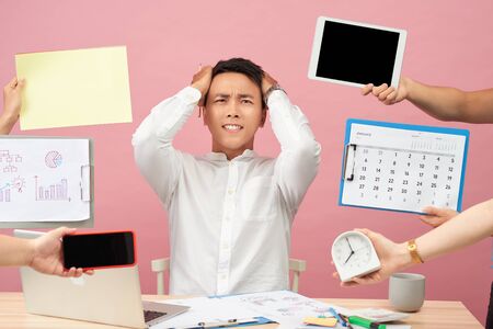 Sad young man sits at desktop, hands with papers, alarm clock, touchpad, notepad with stickers, studies documentation, isolated over pink backgroundの写真素材