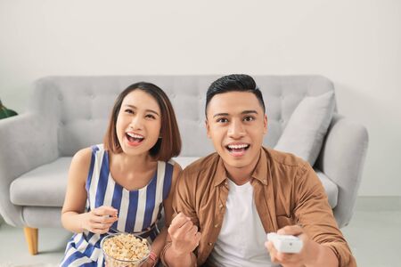 Portrait of a laughing young Asian couple hugging while sitting together at home and watching TV