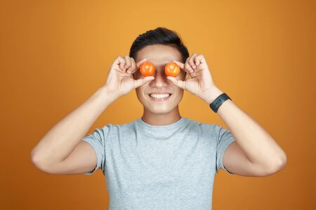 Young Asian man take fun with tomatoes. Isolated on orange backgroundの写真素材