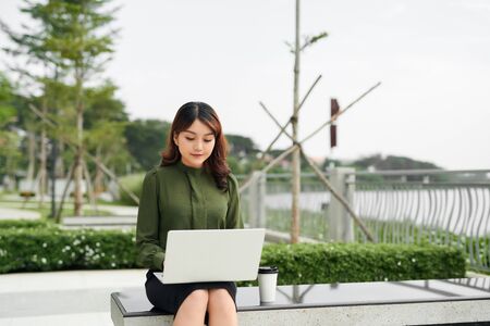 Portrait of busy sales woman sitting at business park and using her laptop while writing text on mobile. Businesswoman working online.の写真素材