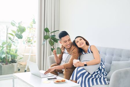 Happy young Asian Couple At Home Using Laptop Computer when sitting on sofaの写真素材