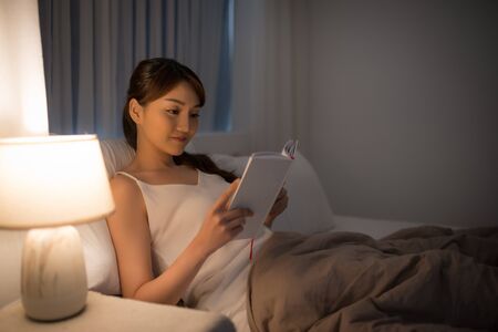 leisure and people concept - young woman reading book in bed at night homeの写真素材