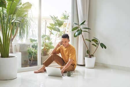 Guy Talking On Cellphone Using Laptop Sitting On Floor At Homeの写真素材