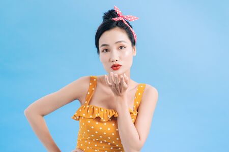 Cute lovely young woman in yellow dress with white polka dots standing and sending kiss over blue backgroundの写真素材