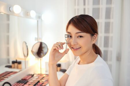 Portrait of beautiful young Asian woman looking at the mirror using eyelash curlerの写真素材