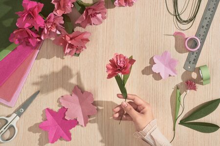 Girl hand holding paper flower over wood background with color paper flowers, ruler, scissors. top view, flatlayの写真素材