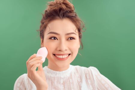 Beauty portrait of a beautiful smiling young Vietnamese woman standing isolated over green background, holding makeup spongeの写真素材
