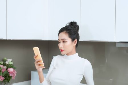Portrait of beautiful Asian woman using phone in kitchen.の写真素材