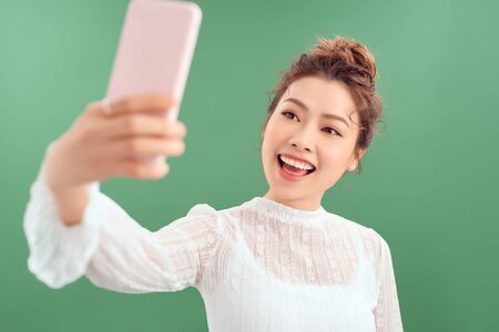 Portrait of cool cheerful girl having video-call holding smart phone in hand shooting selfie on front camera isolated on green backgroundの写真素材