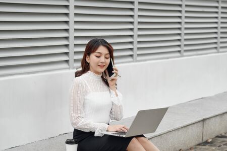 Smiling asian girl sitting on stairs and using a laptop. Asian business woman using laptop computer and talking on her cell smartphoneの写真素材