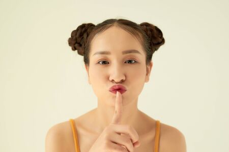 Keep in secret! Portrait of playful positive Asian teen girl with buns hairstyle showing silence gesture over light background.の写真素材