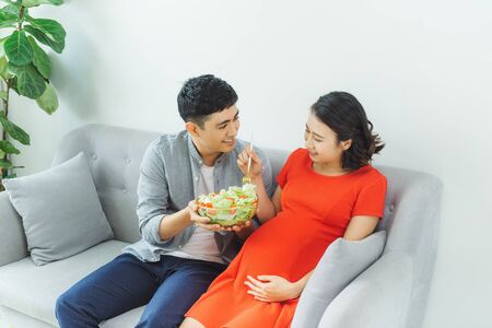 Lovely young pregnant couple relaxing on a couch together, woman having healthy breakfastの写真素材