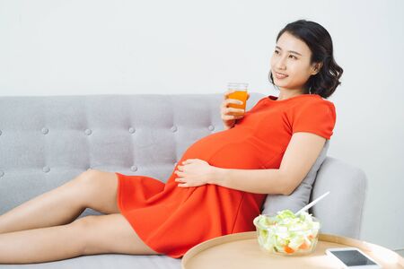 Pretty young Asian pregnant woman eating salad and drinking milk when sitting on sofa.の写真素材