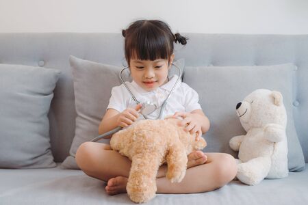 Pretty little girl wearing stethoscope, sitting on the sofa, looking after a sick teddy-bear.の写真素材
