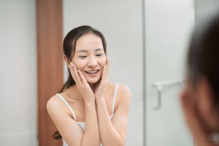 Beauty skin care. Young beautiful teenage girl touching her face before the mirror, enjoying her clean skinの写真素材
