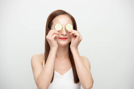 Young beautiful asian lady holding slices of cucumber near her face and smiling isolated over white background.の写真素材