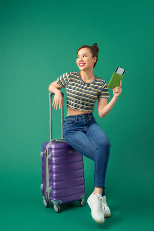 Joyful young Asian girl sitting on suitcase and holding passport, flight ticket to travel over green background.の写真素材