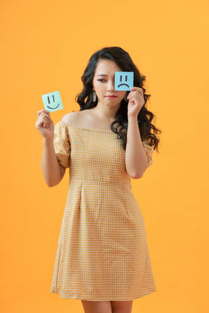 Emotional woman holding note paper with drawn emoticons on yellow backgroundの写真素材