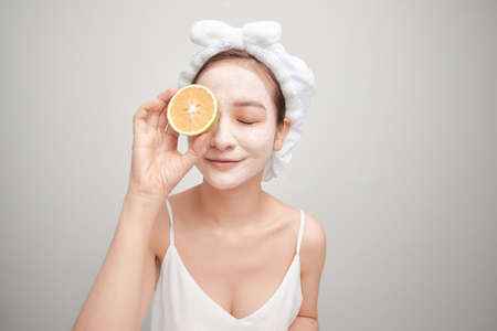 Young woman with facial clay mask holding orange fruit slice covering eyesの写真素材