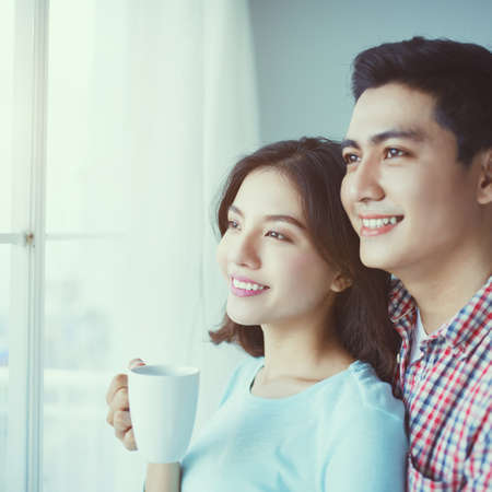 Young Asian couple embracing behind the window and drinking coffee.の写真素材