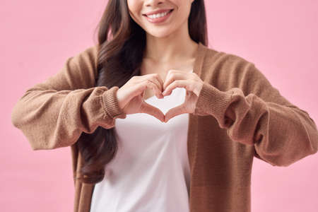smiling young asian woman showing heart gesture with two hands and looking at camera isolated over...