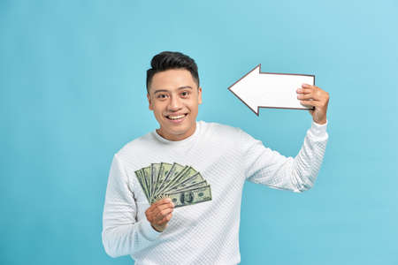 Joyful young business man holding arrow lots bunch of dollars banknotes cash money isolated on blue...