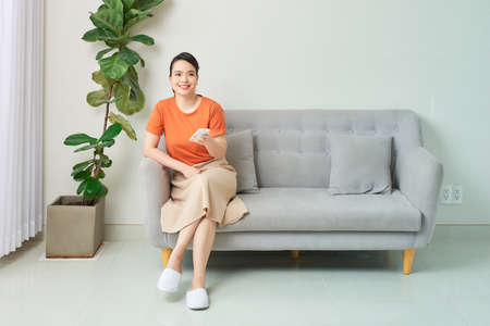 Smiling beautiful woman using air conditioner remote control while sitting on sofa at homeの写真素材