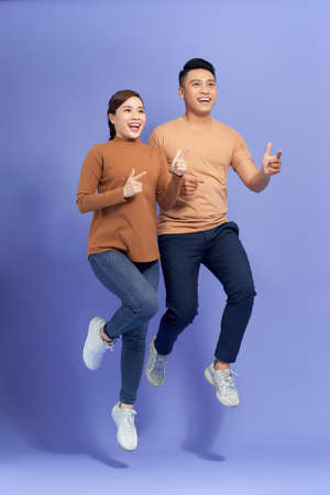 Excited young Asian couple man and woman jumping and pointing with happy and fun over purple...