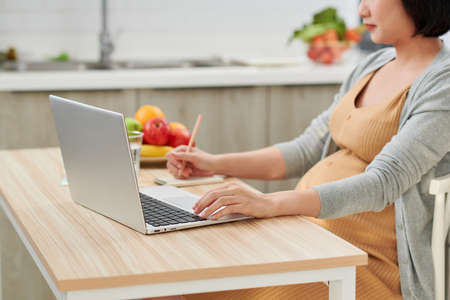 young pregnant woman using laptop while leaning on kitchen counter at homeの写真素材