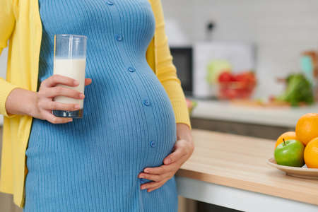 Young pregnant woman with glass of milk standing at kitchenの写真素材