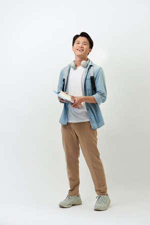 Cheerful young Asian student man with books, headphone, backpack in casual standing over white background.の写真素材