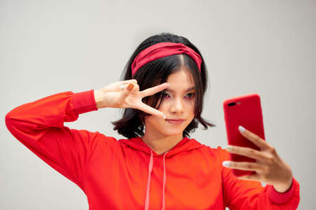 Photo of joyful young woman taking selfie photo on smartphone and gesturing peace sign isolated over gray backgroundの写真素材