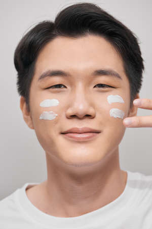 Skin care. Portrait of handsome young man applying cream to his face, isolated on white backgroundの写真素材