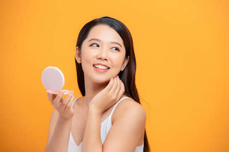 Beauty portrait of a cheerful attractive woman examining her face while looking at the mirror isolated over yellow backgroundの写真素材