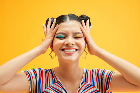 Young woman with colorful eyes smiles and covers her face with her hands, posing on yellow backgroundの写真素材