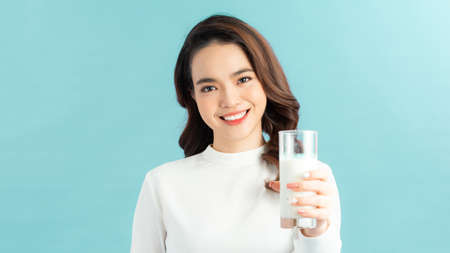 Young beautiful girl holding glass with milk in hand happily looking in camera over blue background