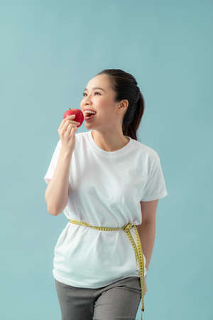 Beautiful sporty woman using tape measure eating apple fruit over blue backgroundの写真素材