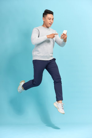 Image of excited young man isolated over blue wall background make selfie by mobile phone jumping.の写真素材